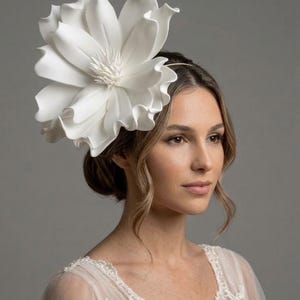 Könnte beinhalten: Ein großer, weißer, skulpturaler Blumen-Kopfschmuck an einem dünnen, metallischen Haarreif. Das Accessoire besteht aus geschichteten, geschwungenen Blütenblättern mit einer detaillierten, stacheligen Mitte in 3D-Optik.