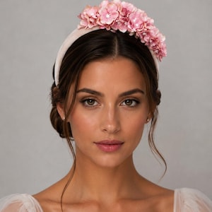 Op de afbeelding: Een lichtroze fluwelen haarband versierd met een tros kunstmatige roze hortensiabloemen aan de bovenkant. Het bloemstuk is voorzien van meerdere gelaagde bloemblaadjes in verschillende tinten roze, wat zorgt voor een dicht en gestructureerd accent.