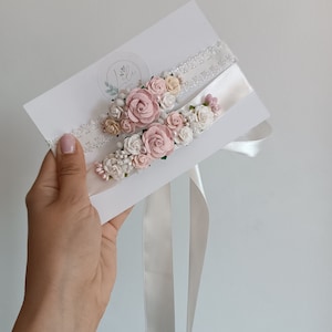 May include: Two white lace and satin garters decorated with clusters of small fabric roses in shades of dusty pink, cream, and white, accented with tiny pearl beads and green leaves, attached to a white display card with a circular logo.