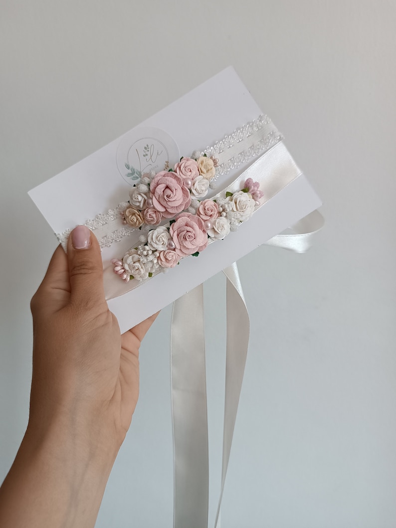May include: Two white lace and satin garters decorated with clusters of small fabric roses in shades of dusty pink, cream, and white, accented with tiny pearl beads and green leaves, attached to a white display card with a circular logo.