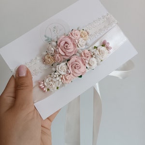 May include: Two white wedding garters featuring floral embellishments in shades of dusty pink, cream, and white. The garters are decorated with small fabric roses, tiny pearl-like beads, and green leaf accents, attached to white lace and satin bands.