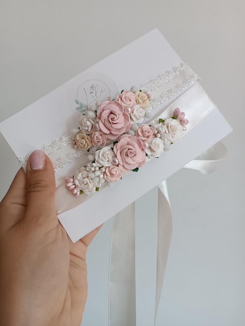 May include: Two white wedding garters featuring floral embellishments in shades of dusty pink, cream, and white. The garters are decorated with small fabric roses, tiny pearl-like beads, and green leaf accents, attached to white lace and satin bands.
