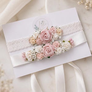 May include: Two leg garters displayed on a white card. The top garter features white lace with a cluster of pink and cream fabric roses and small pearls. The bottom garter is made of smooth white satin with a similar floral arrangement of pink and cream roses, tiny white buds, and pearls.