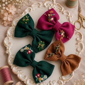 Può includere: Quattro fiocchi per capelli in tessuto verde scuro, magenta e marrone, ciascuno caratterizzato da intricati ricami floreali con piccole perline. I fiocchi sono disposti su un piatto decorativo bianco, con rocchetti di filo e forbici.