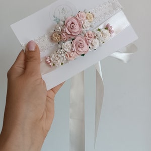 May include: Two white lace and satin garters attached to a white display card. Each garter is decorated with a cluster of small fabric roses in shades of dusty pink, cream, and white, accented with tiny pearl beads and small green leaf details.
