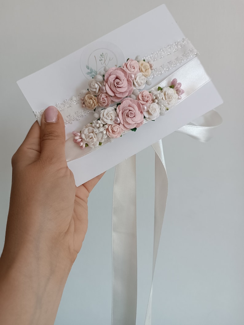 May include: Two white lace and satin garters attached to a white display card. Each garter is decorated with a cluster of small fabric roses in shades of dusty pink, cream, and white, accented with tiny pearl beads and small green leaf details.