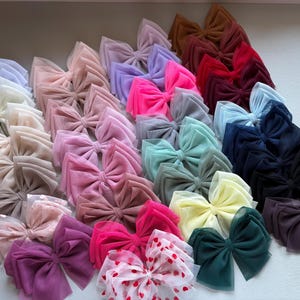 May include: A collection of handmade tulle bows in various colors, including pink, purple, blue, green, and white. The bows are arranged in a semi-circle on a white surface.