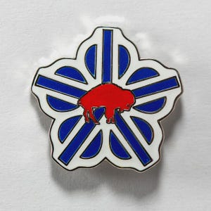 May include: A white, blue, and red enamel pin in the shape of a five-petal flower. The pin features a red bison in the center, surrounded by blue and white geometric shapes. The pin has a silver border.