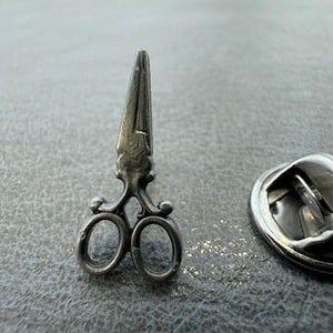 May include: A silver-colored metal pin shaped like a pair of scissors. The pin has a detailed design, with the scissor blades closed and the handles featuring decorative loops. The pin is shown next to a silver-colored pin back.