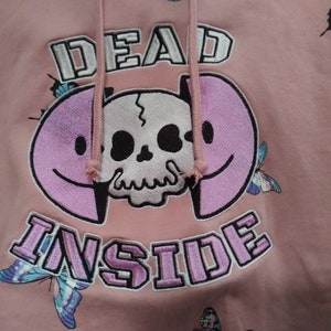 May include: Pink hoodie with a skull and smiley face design. The text "DEAD INSIDE" is embroidered on the front of the hoodie.