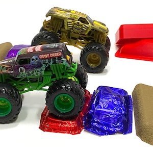 May include: Two toy monster trucks, one green and one gold, are on a track with ramps and jumps. The green truck is labeled "Grave Digger".