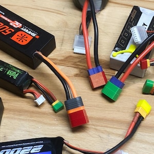 May include: Assortment of black and orange RC batteries and connectors. The batteries have labels with specifications like "5000 mAh" and "2200 mAh". Various colored connectors are attached to the batteries with red, black, and orange wires.