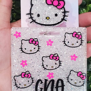 Puede incluir: Un portagafete blanco con purpurina con un diseño de Hello Kitty rosa y el nombre "GINA" impreso en negro. El portagafete está unido a una cadena plateada.