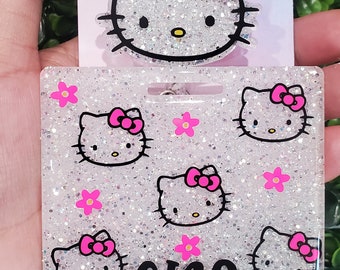 Può includere: Un porta badge bianco con glitter con un design Hello Kitty rosa e il nome "GINA" stampato in nero. Il porta badge è attaccato a una catena argentata.
