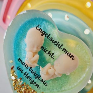 Könnte beinhalten: Ein herzförmiger Papiergewicht aus Harz mit einem blauen und grünen Farbverlauf. Zwei kleine weiße Babyfiguren befinden sich im Papiergewicht. Der Text "Engel sieht man nicht, man trägt sie im Herzen." ist auf das Papiergewicht gedruckt.