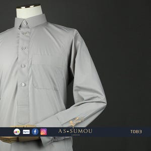 May include: A light gray long-sleeved shirt with a button-up collar and a pocket on the left side. The shirt has a traditional design and is made of a smooth, wrinkle-resistant fabric. The shirt is labeled "AS-SUMOU LONDON" and "TO83".