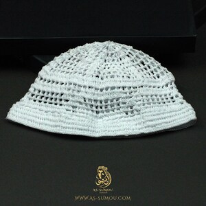May include: A white crocheted skullcap featuring a complex openwork pattern with horizontal bands of dense stitching. The item is displayed above a gold logo and the text AS-SUMOU LONDON WWW.AS-SUMOU.COM printed on a dark surface.