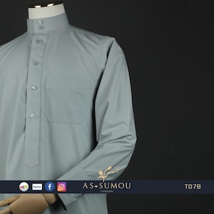 May include: A light blue long-sleeved shirt with a button-up collar and a pocket on the left chest. The shirt has a traditional design and is made of a smooth, lightweight fabric. The shirt has the text "AS-SUMOU LONDON" on the cuff and "TD78" on the bottom right corner.
