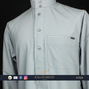 May include: A light gray long-sleeved shirt with a button-up collar and a pocket on the left side. The shirt has a decorative embroidered design on the right side. The text "AS-SUMOU LONDON" and "KT69" are visible on the bottom of the shirt.