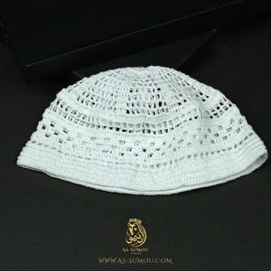 May include: A white crocheted kufi cap featuring a repeating openwork geometric pattern throughout the crown and sides. The skullcap has a solid crocheted band at the base and a circular top. The logo for AS-SUMOU LONDON is printed below.