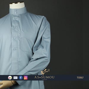 May include: A light blue long-sleeved shirt with a stand-up collar and button closure. The shirt has a single pocket on the left chest and is made of a smooth, wrinkle-resistant fabric. The shirt is displayed on a mannequin against a dark gray background. The brand name "AS-SUMOU LONDON" is printed on the bottom right corner of the image, along with the product code "T082".