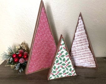 May include: Three wooden Christmas tree ornaments, each with a different patterned paper covering. The largest tree is red with white text that says "Merry Christmas". The middle tree is green with a red and white holly pattern. The smallest tree is white with black musical notes.