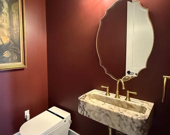 May include: A powder room with deep red walls featuring a white smart toilet, a wall-mounted marble sink with gold faucets, a scalloped gold-framed mirror, a three-light glass fixture, and a framed painting of a person holding a fan in a gold frame.