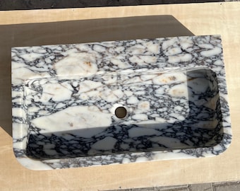 May include: A white marble sink with black and brown veining. The sink has a rounded rectangular shape and a single drain hole.