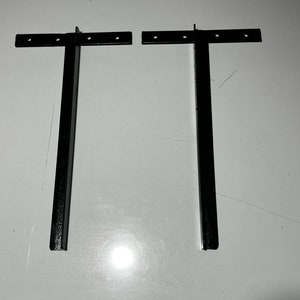 May include: Two black metal brackets with holes for mounting. The brackets are in the shape of a T, with a long vertical leg and a short horizontal leg.