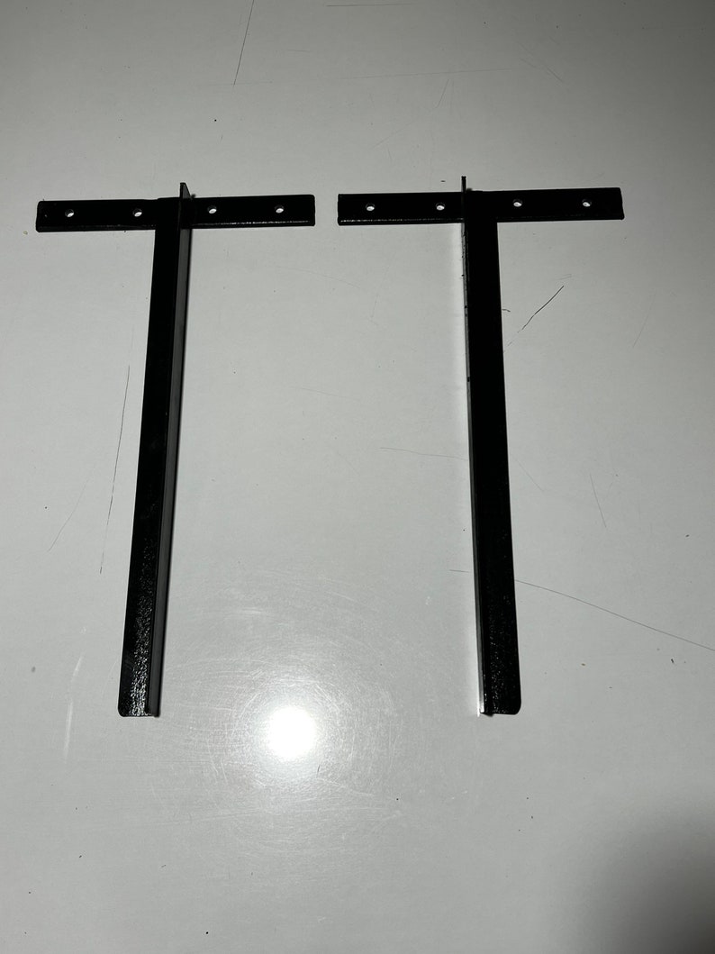 May include: Two black metal brackets with holes for mounting. The brackets are in the shape of a T, with a long vertical leg and a short horizontal leg.