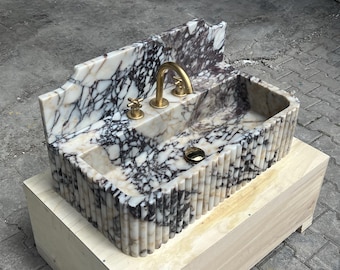 May include: A rectangular marble sink with a white and dark grey veined pattern. The sink has a fluted exterior and gold-coloured tap and fixtures. The sink is on a wooden base.