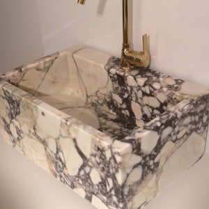 Pu&ograve; includere: Un lavabo in marmo bianco con un rubinetto dorato. Il lavabo ha una forma rettangolare ed &egrave; realizzato in pietra naturale. Il rubinetto &egrave; montato a parete e ha un'unica manopola.