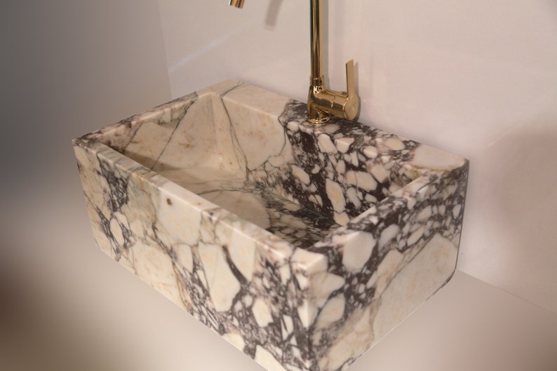 Pu&ograve; includere: Un lavabo in marmo bianco con un rubinetto dorato. Il lavabo ha una forma rettangolare ed &egrave; realizzato in pietra naturale. Il rubinetto &egrave; montato a parete e ha un'unica manopola.