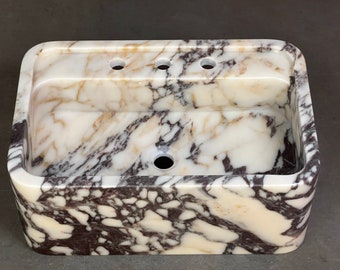 Puede incluir: Un lavabo de mármol blanco con vetas marrones y moradas. El lavabo tiene una forma rectangular con bordes redondeados y tres agujeros en el borde superior. El lavabo está hecho de piedra natural y tiene un aspecto único y artesanal.