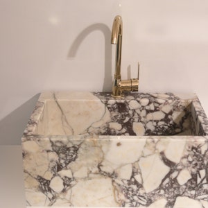 Pu&ograve; includere: Un lavabo in marmo bianco con un rubinetto dorato. Il lavabo &egrave; rettangolare ed &egrave; fatto di marmo bianco con venature marroni e grigie.