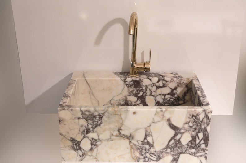 Pu&ograve; includere: Un lavabo in marmo bianco con un rubinetto dorato. Il lavabo &egrave; rettangolare ed &egrave; fatto di marmo bianco con venature marroni e grigie.