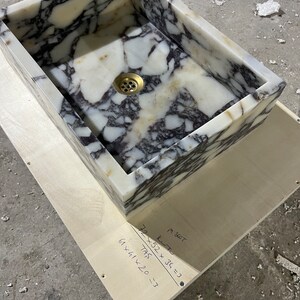 Peut inclure: Un &eacute;vier en marbre blanc avec des veines grises. L'&eacute;vier est rectangulaire et poss&egrave;de un drain au centre. L'&eacute;vier est pos&eacute; sur une surface en bois avec des mesures manuscrites : "52 x 36 = 1872", "61 x 41 x 20 = 51420", et "72 / 745 = .3".