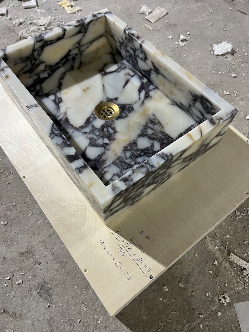 Peut inclure: Un &eacute;vier en marbre blanc avec des veines grises. L'&eacute;vier est rectangulaire et poss&egrave;de un drain au centre. L'&eacute;vier est pos&eacute; sur une surface en bois avec des mesures manuscrites : "52 x 36 = 1872", "61 x 41 x 20 = 51420", et "72 / 745 = .3".