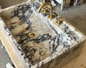 May include: Marble sink with gold taps and fittings. The sink has a rectangular basin with a white and beige background and dark grey veining. The gold-coloured tap is curved.