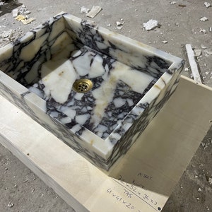 Peut inclure: Un &eacute;vier rectangulaire en marbre blanc avec des veines noires. L'&eacute;vier est dot&eacute; d'un drain en laiton. L'&eacute;vier est pos&eacute; sur une base en bois avec des mesures manuscrites en pouces.