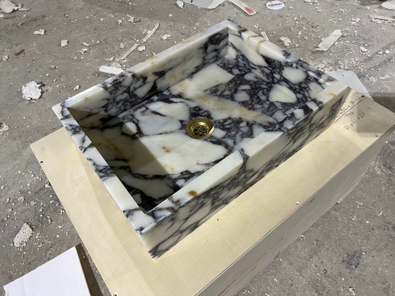 Peut inclure: Un &eacute;vier en marbre blanc avec des veines grises et violettes. L'&eacute;vier est rectangulaire et poss&egrave;de un drain en laiton. L'&eacute;vier est pos&eacute; sur une base en bois.