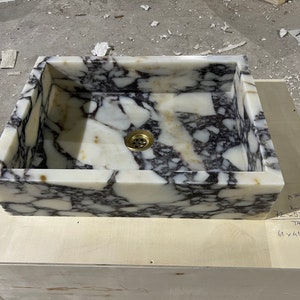 Peut inclure: Un &eacute;vier en marbre blanc avec des veines noires. L'&eacute;vier est rectangulaire et poss&egrave;de un drain au centre. L'&eacute;vier est pos&eacute; sur une surface en bois.