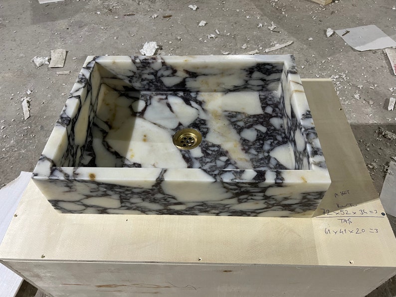 Peut inclure: Un &eacute;vier en marbre blanc avec des veines noires. L'&eacute;vier est rectangulaire et poss&egrave;de un drain au centre. L'&eacute;vier est pos&eacute; sur une surface en bois.