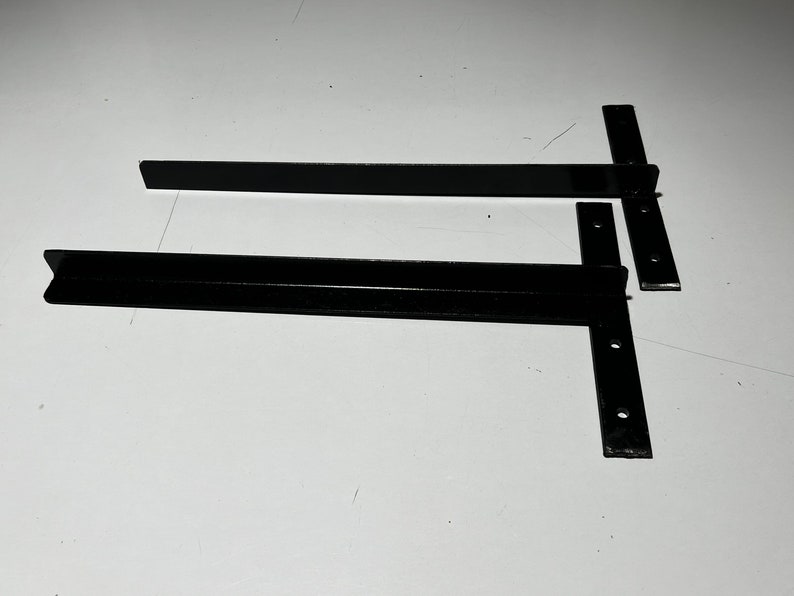 May include: Two black metal shelf brackets with a flat and angled design. The brackets have multiple holes for mounting.
