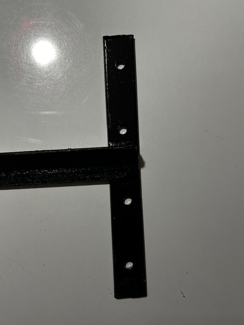 May include: Black metal bracket with four holes for mounting. The bracket has a T-shape with a long horizontal arm and a shorter vertical arm.