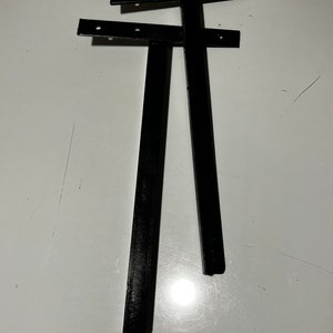 May include: Two black metal brackets with holes for screws. The brackets are L-shaped and have a flat surface on one side.