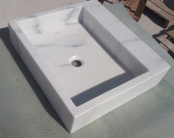 May include: A white marble bathroom sink with a rectangular shape and a single drain hole. The sink is sitting on a gray textured surface.