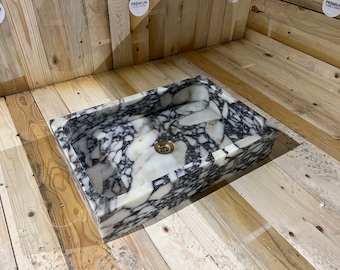 May include: A white marble sink with black veining. The sink is rectangular and has a drain in the center. The sink is sitting on a wooden surface.