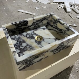 Peut inclure: Un &eacute;vier rectangulaire en marbre blanc avec un drain en laiton. L'&eacute;vier est pos&eacute; sur un meuble en bois.
