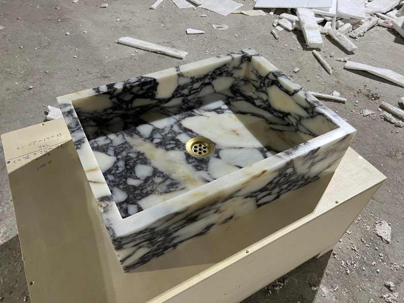 Peut inclure: Un &eacute;vier rectangulaire en marbre blanc avec un drain en laiton. L'&eacute;vier est pos&eacute; sur un meuble en bois.